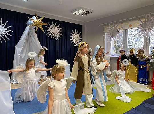 Józef i Maryja z aniołkami które trzymają białe flagi a pod oknem stoją Trzej Królowie