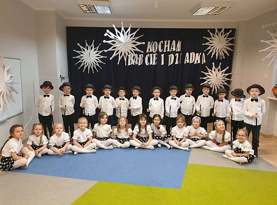 Grupa dzieci na tle dekoracji, chłopcy z kapeluszami i laseczkami, dziewczynki siedzą przednimi