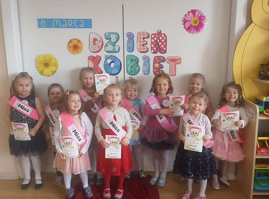 Dziewczynki stoją w szarfach z napisem Miss oraz z dyplomami